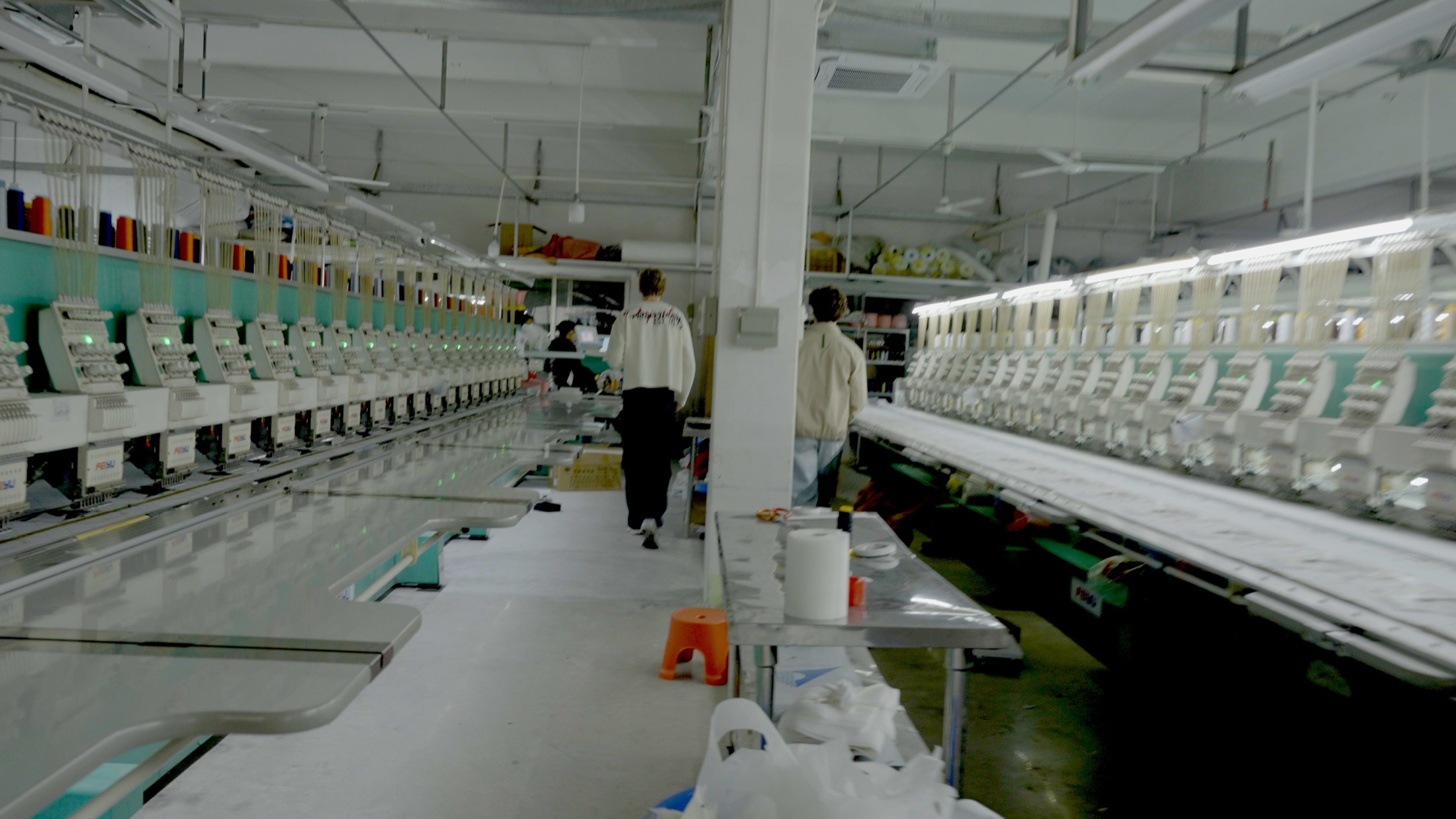 A long machine hall in a partner factory, two people walking the centre aisle between the rows